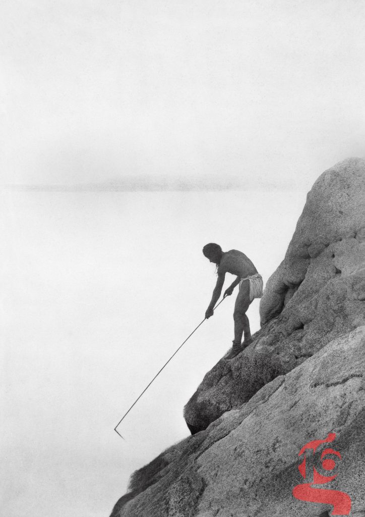 Homme paviotso pêchant à la gaffe, penché sur un rocher au-dessus d'une eau calme, photographié par Edward S. Curtis vers 1924.