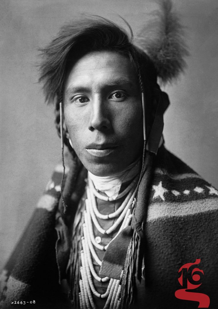 Portrait frontal d’un homme aaniiih portant une coiffure ornée d’une plume et de longues parures, photographié par Edward S. Curtis en 1908.