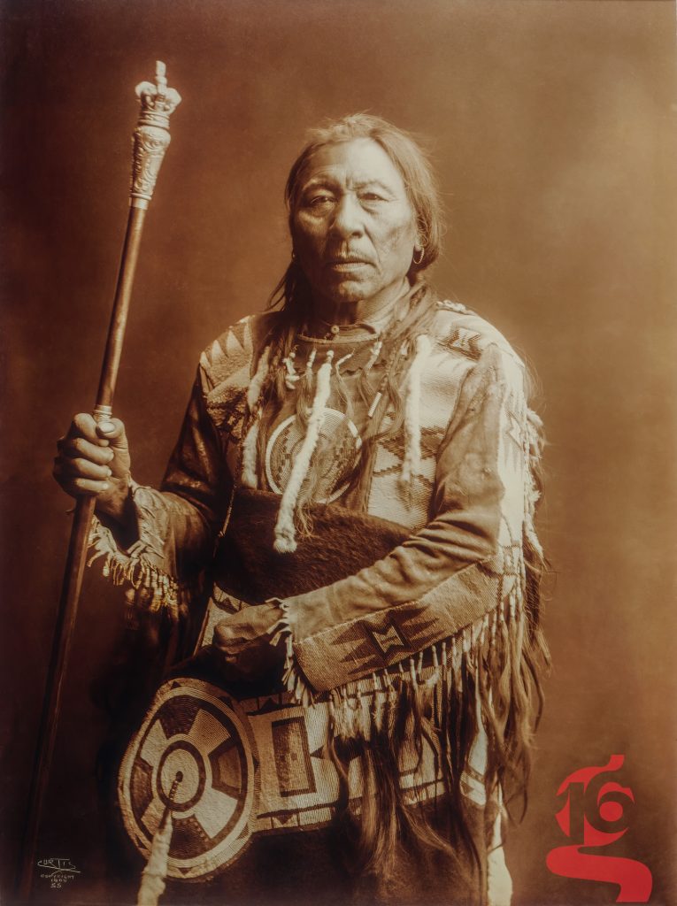 Portrait de Medicine Crow, homme Apsáalooke tenant un bâton orné, photographié par Edward S. Curtis en 1908.