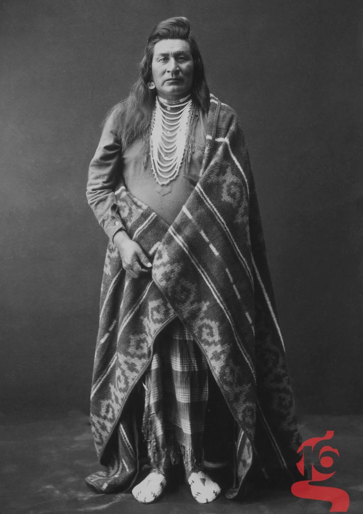 Portrait en pied d’un homme nez-percé portant couverture et colliers, photographié par Edward S. Curtis vers 1899.