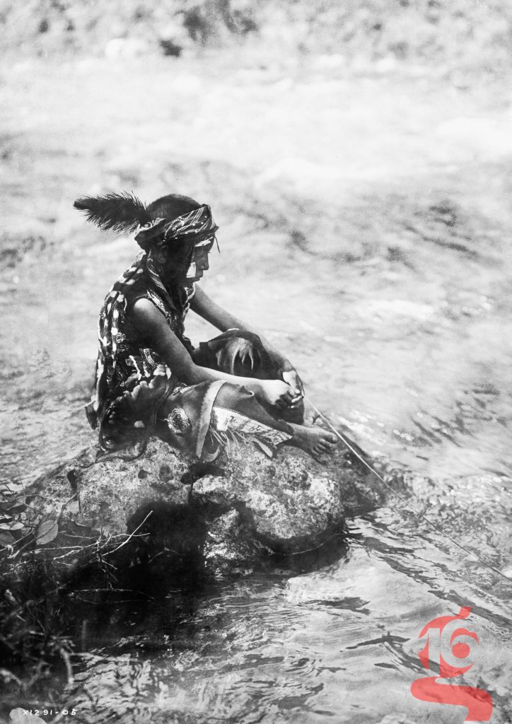 Jeune garçon apsáalooke assis sur un rocher pendant qu’il pêche dans une rivière, photographié par Edward S. Curtis en 1905.