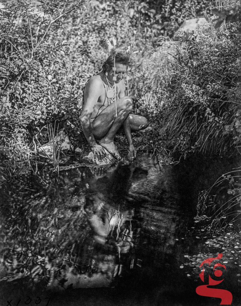 Homme accroupi au bord d’un ruisseau, dont le reflet apparaît dans l’eau, photographié par Edward S. Curtis en 1905.