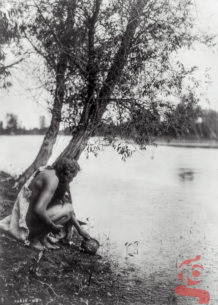 Homme hidatsa accroupi au bord d’un cours d’eau avec un récipient, photographié par Edward S. Curtis vers 1908.