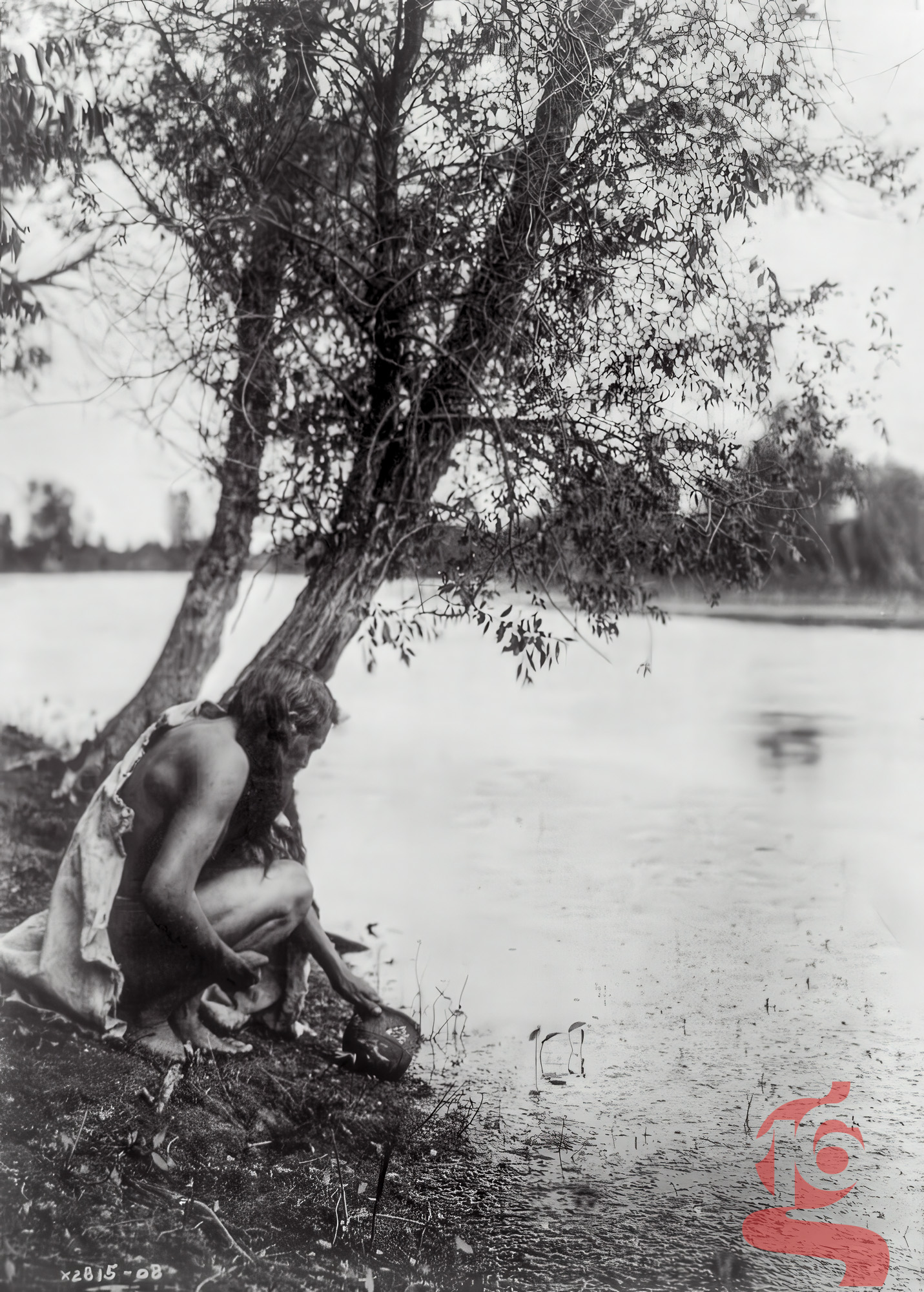 Homme hidatsa accroupi au bord d’un cours d’eau avec un récipient, photographié par Edward S. Curtis vers 1908.