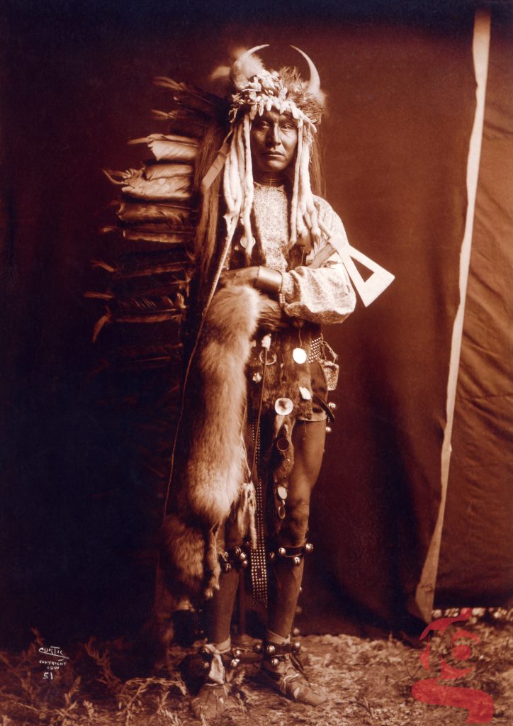Portrait en pied d’Iron Breast, homme Piegan en tenue cérémonielle ornée de plumes, fourrure et cornes, photographié par Edward S. Curtis en 1900.
